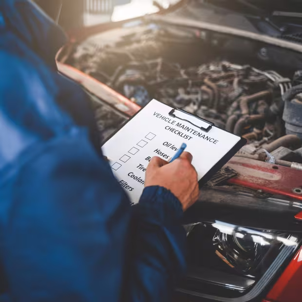 Mechanic noting findings on a vehicle maintenance checklist over an open engine bay